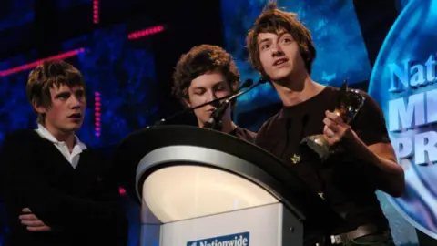 PA Media An image of three members of the Arctic Monkeys at an awards show accepting a trophy 
