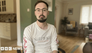 Darren is wearing a long sleeve white t-shirt and is sat in a wheelchair in his home. He has dark hair and facial hair and is looking into the camera.