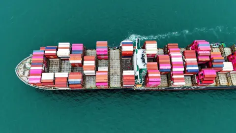 CFOTO/Getty Images A large cargo ship staked with pink, blue and white container ships sails from right to left through the picture, a small trail of white foam in the picture. 