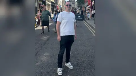 Handout Martin Fletcher is standing in a street and is smiling while wearing sunglasses, a white top and dark trousers.
