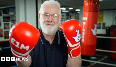 Peter Rogers smiles triumphantly as he poses in a boxing ring with red boxing gloves on which say £1m and have the National Lottery logo. Gold confetti falls around him. He has short white hair and short white beard. He is wearing glasses and a blue polo top.