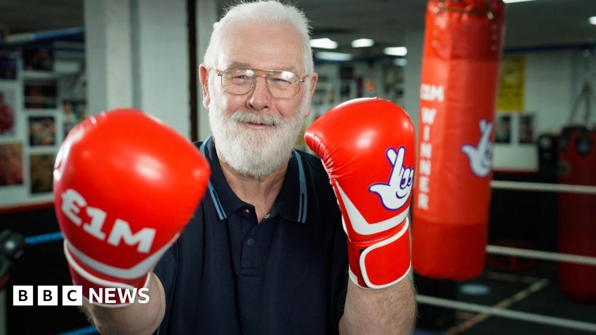 Peter Rogers smiles triumphantly as he poses in a boxing ring with red boxing gloves on which say £1m and have the National Lottery logo. Gold confetti falls around him. He has short white hair and short white beard. He is wearing glasses and a blue polo top.