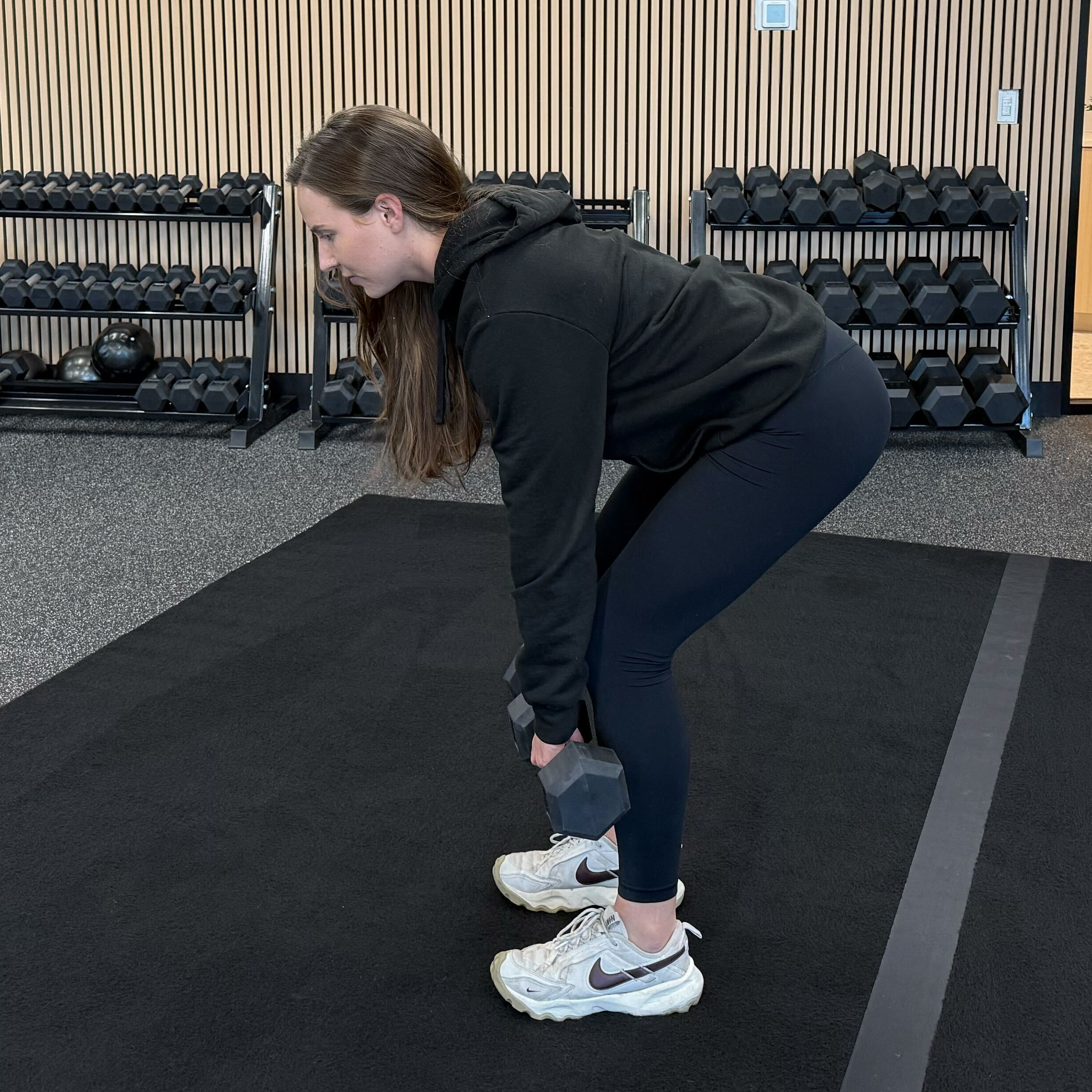Felicia Hernandez, a personal trainer for Eden Health Club, performs a Romanian deadlift with dumbbells. She is standing with feet shoulder-width apart, hinging forward at the hips, dumbbells held at her shins.