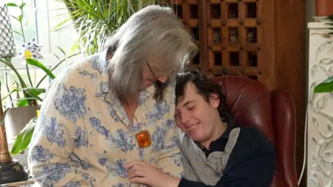 Richard Knights/BBC Chantal Chaervey rests on the arm of red leather chair which her son Harry sits in. She has shoulder length grey hair and wears a white blouse with blue flowers on it. Harry smiles as he rests his head on his mother's shoulder. He has dark hair and wears a grey and blue long sleeved top. 