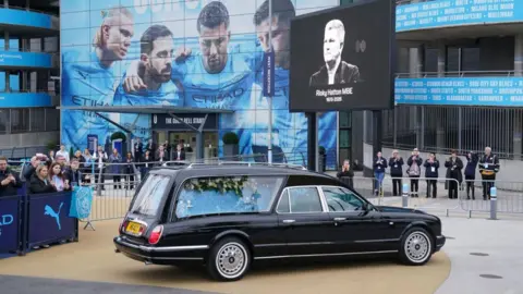 PA The black hearse is parked up outside The Etihad Stadium. A black and white photograph of Hatton is on the screen in front of the stadium.