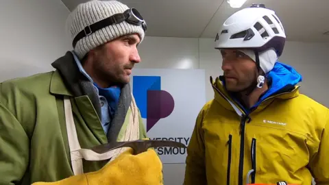 Hugo and Ross Turner in a cold chamber at the University of Portsmouth. One brother in old clothes and one in new clothes. Both sets of clothing are bulky and they both have hats on.