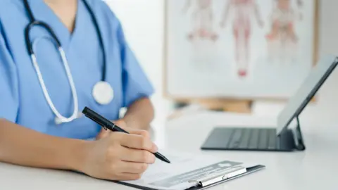 Getty Images A nurse is writing on a clipboard with a black pen. There is a laptop in front of the nurse. On the table is a laptop. The nurse has blue overalls on. The background is blurred but there is a chart on display. 