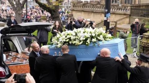 PA Media A blue coffin, which is adorned with white flowers, is carried out of Manchester Cathedral by Ricky Hatton's family and put into the back of a hearse.  Crowds of people can be seen paying their respects in the background. 