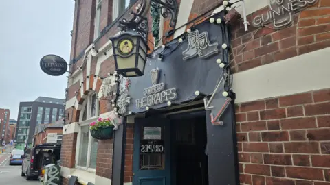Hayley Coyle/BBC Exterior of 'The Grapes' pub, a red-brick building with white trim. A black sign with white lettering and a pint glass graphic reads 'Guinness' and 'The Grapes.' The entrance features a blue door, hanging flower pots, string lights, and multiple Guinness signs, creating a welcoming pub atmosphere.