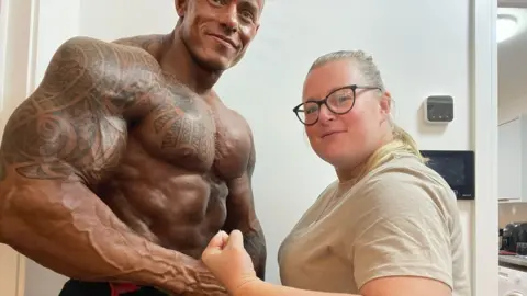 Jay Baggaley Jay Baggaley is smiling and posing while standing next to his partner. She is smiling with her fist clenched while looking at the camera. She has long blonde hair and is wearing a cream coloured T-shirt and glasses. 