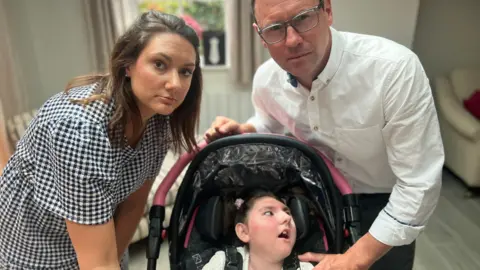 Charlotte, Darcey and Stephen from left to right looking at the camera. Charlotte has shoulder-length brunette hair and is wearing a white and black chequered dress. Darcey is in a black pram with a pink handle. She has dark hair which is bunched up. Stephen, who has glasses is stood behind her, wearing a white shirt.