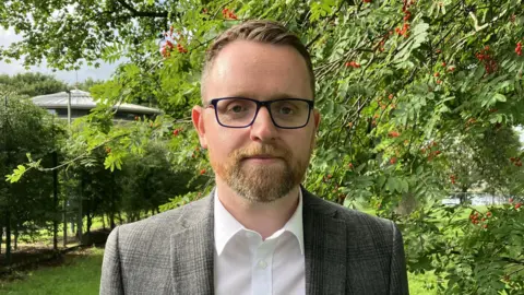 Gary Middleton is wearing a white shirt and a grey blazer, he has black glasses on. He has short head hair and facial hair. Behind him are trees with red berries on them. A building is also visible behind the trees. 