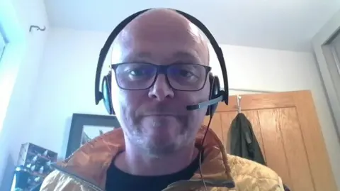 David Tosh is bald with black framed glasses and a light beard. He is wearing a dark top, mustard coloured body warmer and headphones