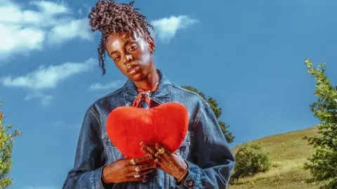 Sony Music Cat Burns holds up a cushion in the shape of a heart, while standing against a countryside landscape, in a promotional photo for her new album How To Be Human