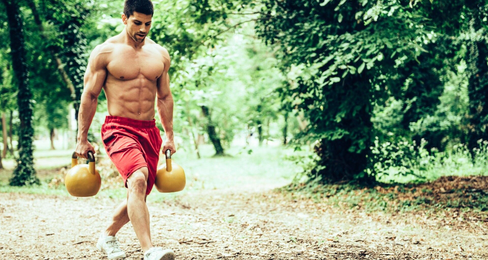 Man walking through a park holding two kettlebells performing a farmer's walk