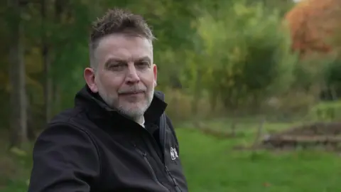 Andy Alcroft/BBC Dom wears a black fleece jacket with the Wildlife Trusts log while sitting on a bench in a field, surrounded by trees
