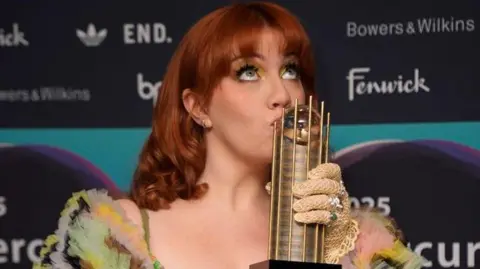 PA Media CMAT kissing a gold trophy. She has dark ginger hair with a fringe and is looking up. She is wearing yellow eyeshadow, yellow fishnet gloves and a multi-coloured top. She is standing in front of a Mercury Prize board which has the names of the awards' sponsors on it.