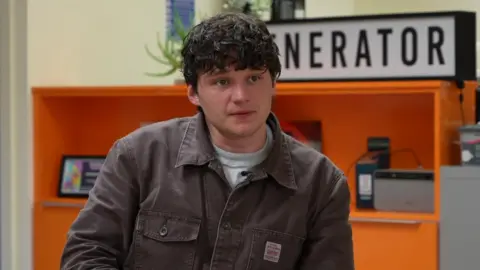 Finn Forster sitting inside Generator studios. He is wearing a brown overshirt and he has brown curly hair.
