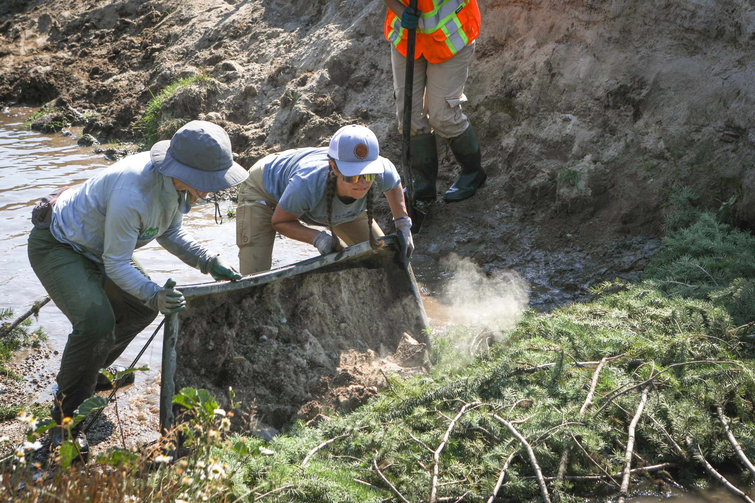 Two women dump a container of dirt onto the bank of a creek's edge.