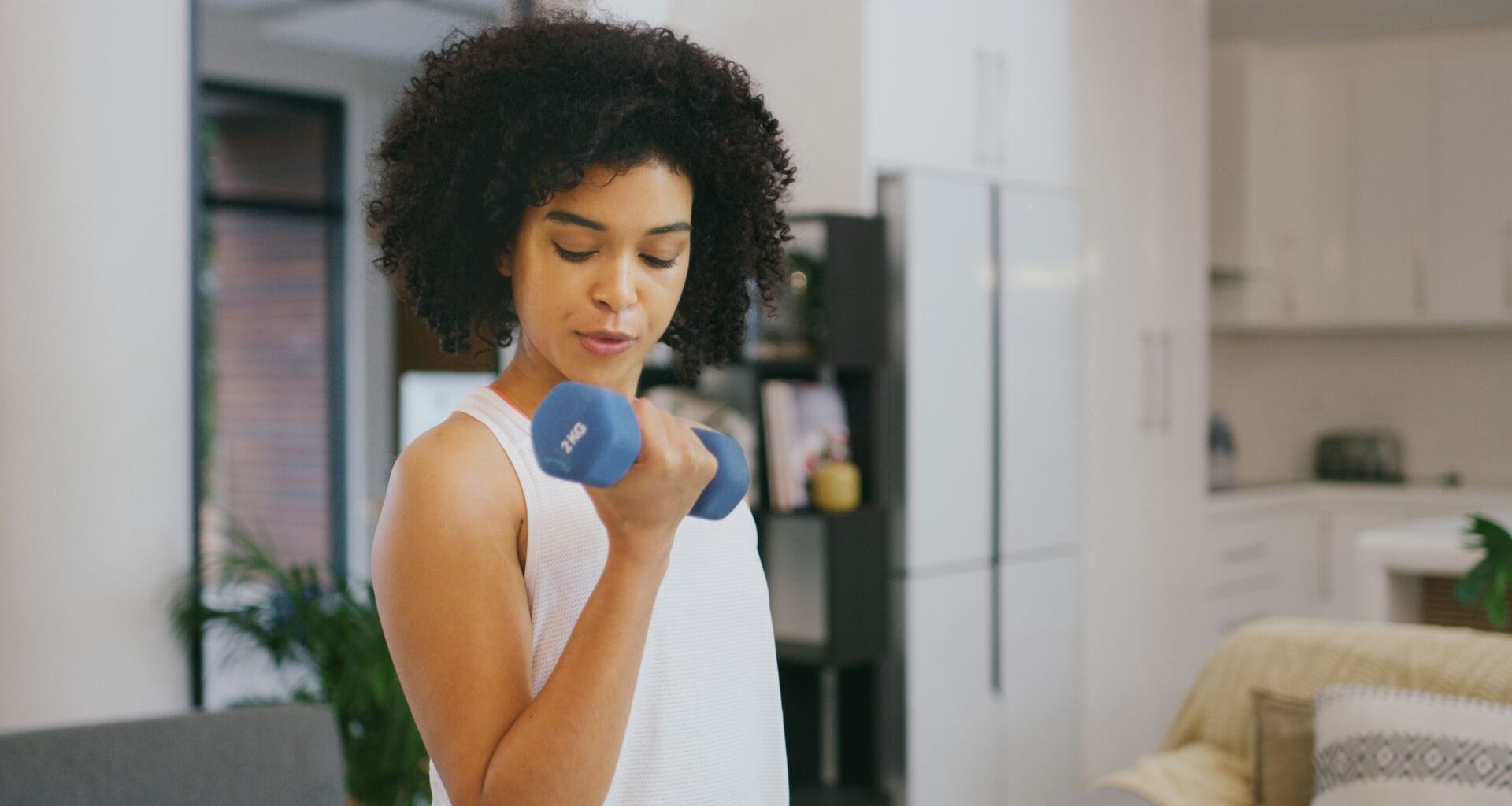 woman curling a blue dumbbell to her biceps, wearing a white vest in a home setting