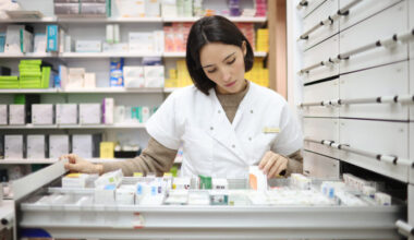 A woman is standing behind a drawer filled with prescription medicines. She is wearing a white lab coat.