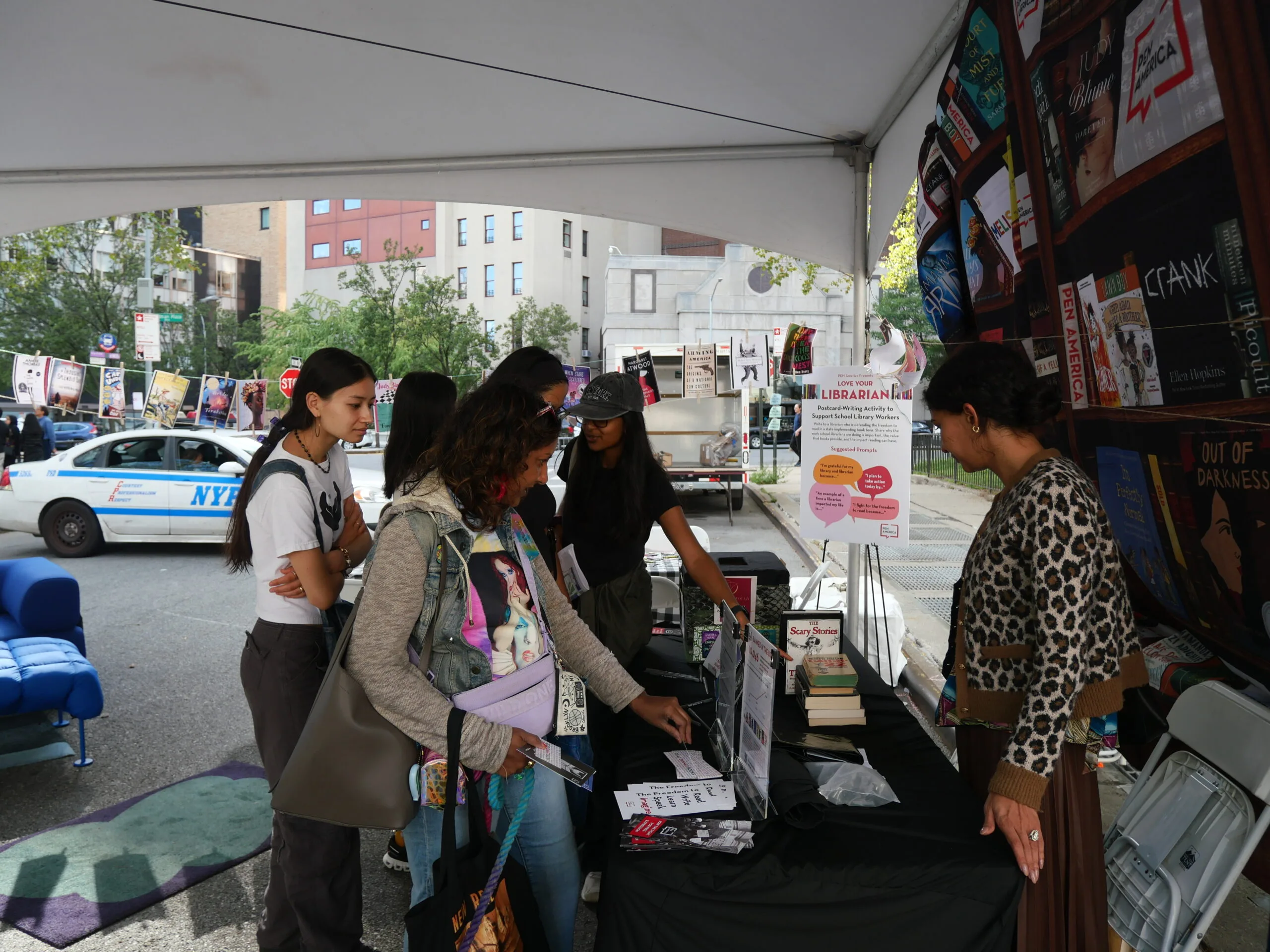 Several people gather under a tent at an outdoor event, examining books and materials on a table. A woman stands behind the table assisting them. A police car and buildings are visible in the background.