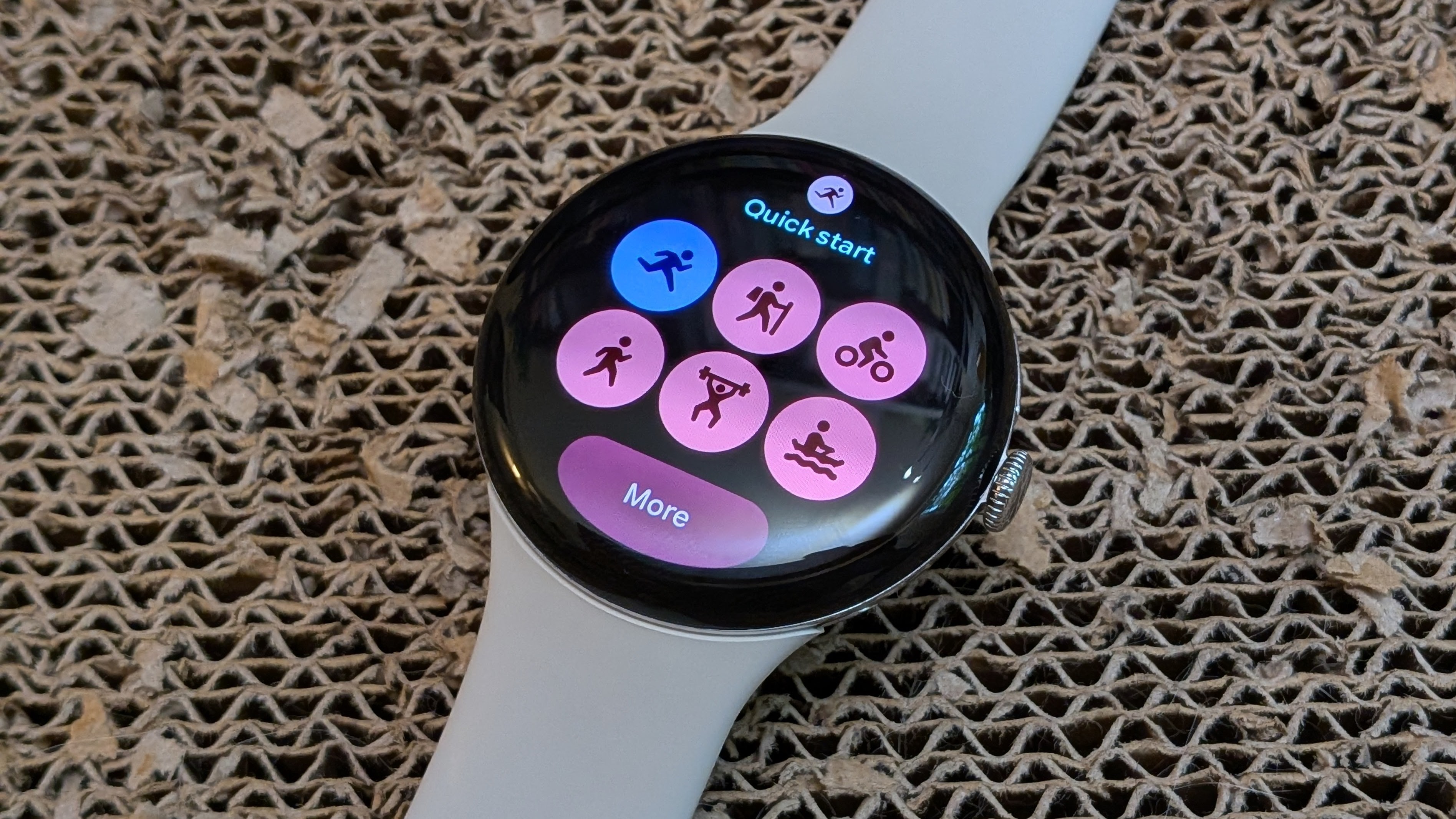 A Pixel Watch 4 sitting on a scratch pad showing the Fitbit Quick Start Tile with pink theming for the buttons