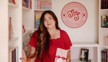 Christina Duran, in a red dress, stands in a bookstore holding a wrapped book. Shelves of books and a wall sign reading “Sugar And Spice Book Co” are visible in the background.