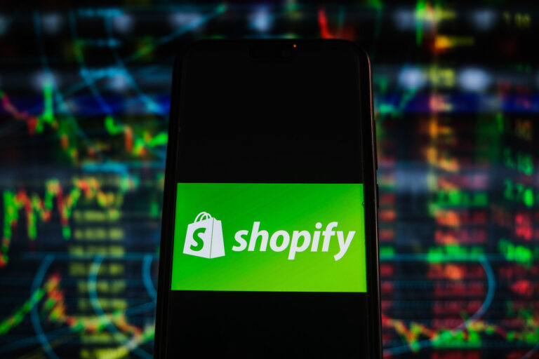 Shopify's logo appears on a smartphone with stock market data on screens in the background.