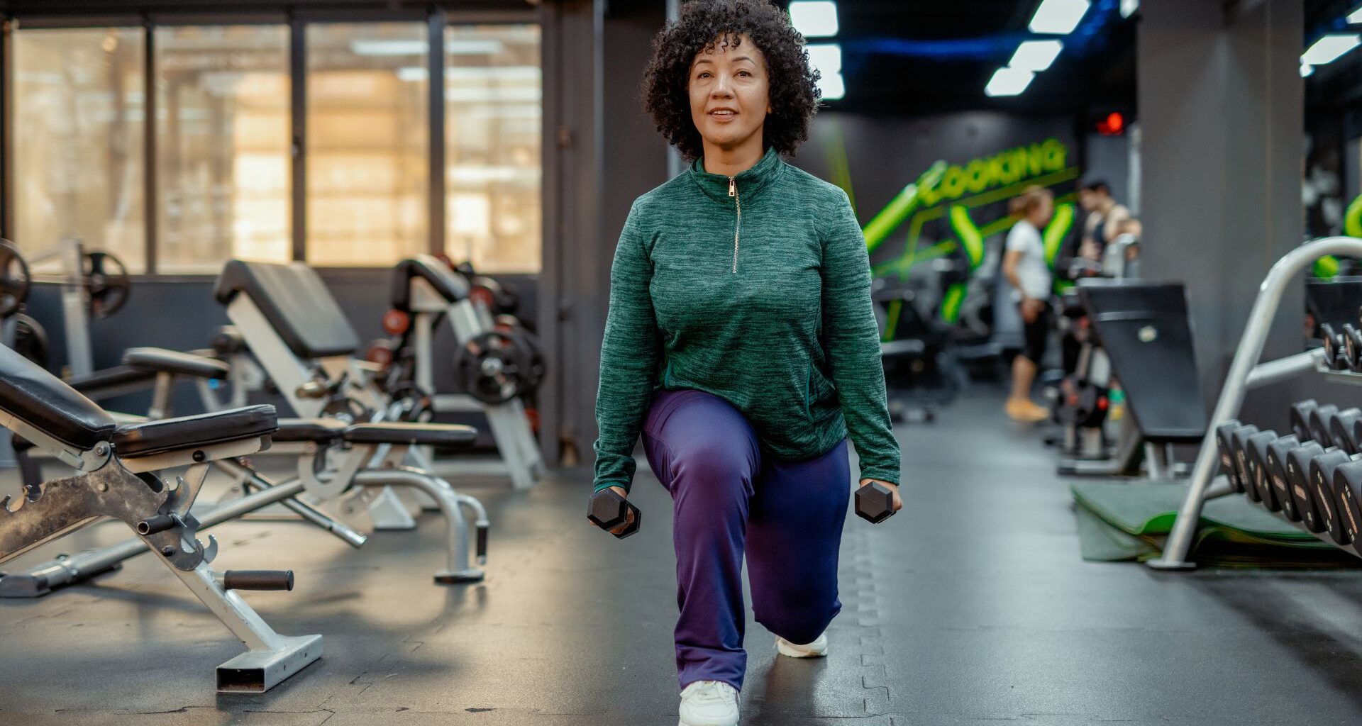 Woman doing reverse lunge, one of the best exercises for 60 and over in the gym holding dumbbells and wearing activewear