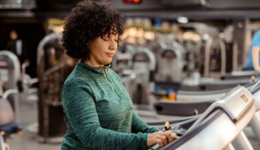 woman in a green long sleeved top on a treadmill and adjusting the settings; the camera shot is from the waist up with a gym setting behind her.