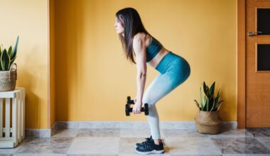 woman in leggings and crop top holding two dumbbells half way through a deadlift. she's sideways to the camera with a yellow wall and a couple of plants behind her