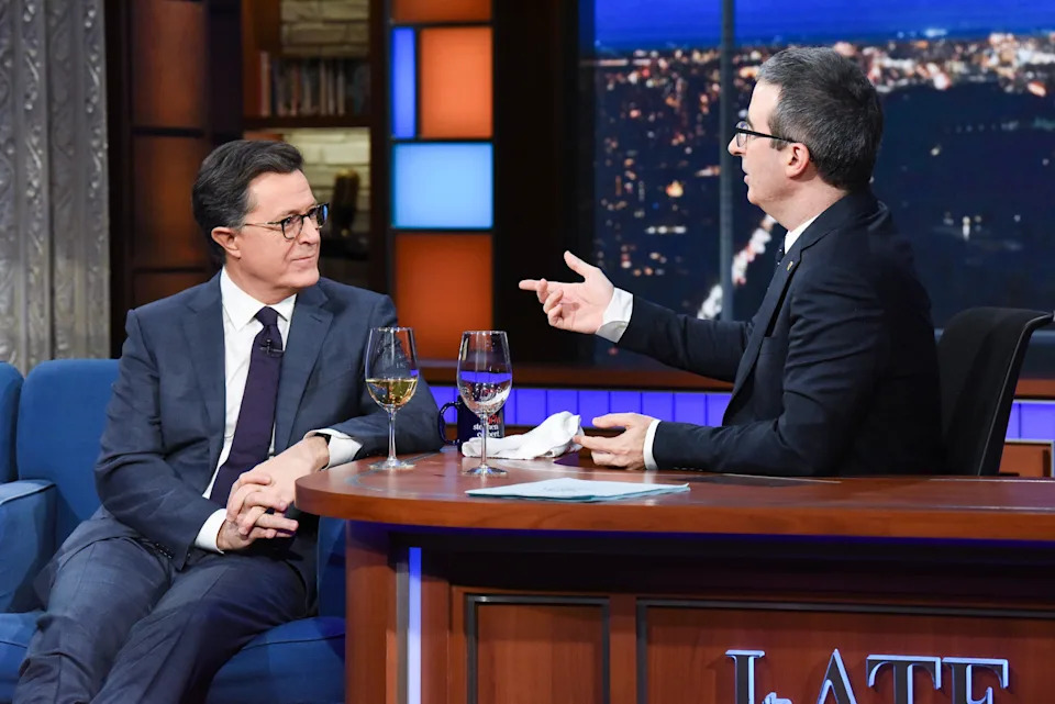 Stephen Colbert and John Oliver have a lively conversation on a late-night talk show set, both seated with wine glasses on the desk
