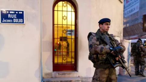 Kenzo Tribouillard/AFP A French soldier standing guard outside a synagogue in a Paris suburb. He is dressed in military fatigues and is carrying a rifle.