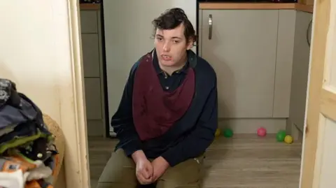 Richard Knights/BBC Harry kneels on the floor within a door frame. He looks away from the camera. He wears a blue jumper with green trousers and a red bib.