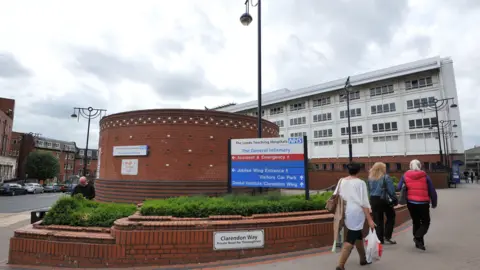 PA Media A general view of Leeds General Infirmary hospital. Members of the public walk up the path to the main building. A blue and white NHS sign stands in the foreground.