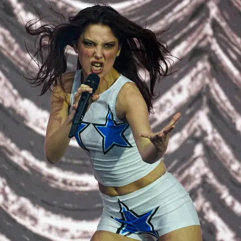 PA Media Ellie Rowsell of Wolf Alice plays at the 2025 Glastonbury festival. She is reaching an arm towards the audience while her hair blows in the wind.