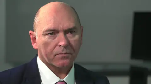 Sir Jim Mackey wearing a white shirt, blue blazer and pink and blue striped tie