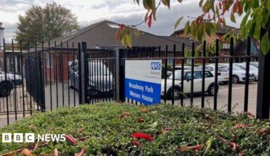A dark-coloured metal fence behind some bushes, with "Broadway Park Wessex House" written on it in white letters on a blue background. The sign also has the NHS logo on it. Behind the fence and sign are parked cars and also a one-story red brick building.