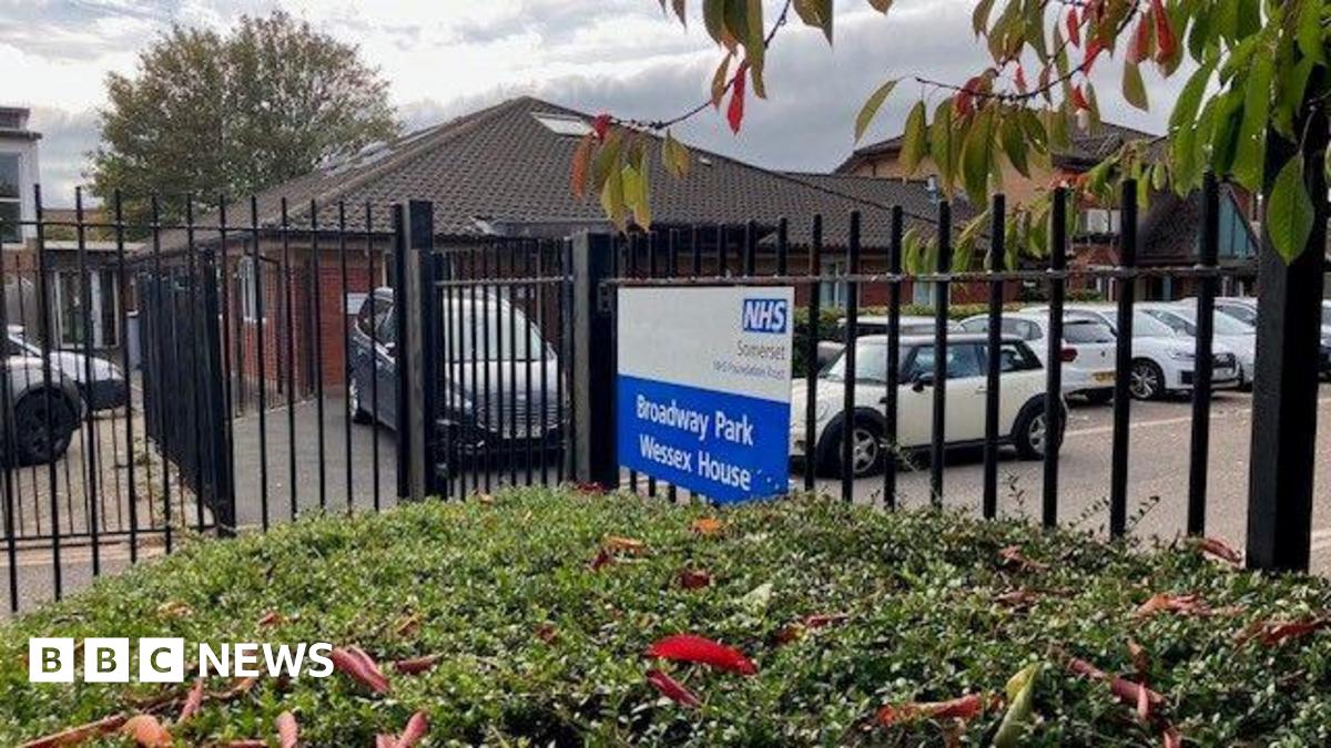 A dark-coloured metal fence behind some bushes, with "Broadway Park Wessex House" written on it in white letters on a blue background. The sign also has the NHS logo on it. Behind the fence and sign are parked cars and also a one-story red brick building.