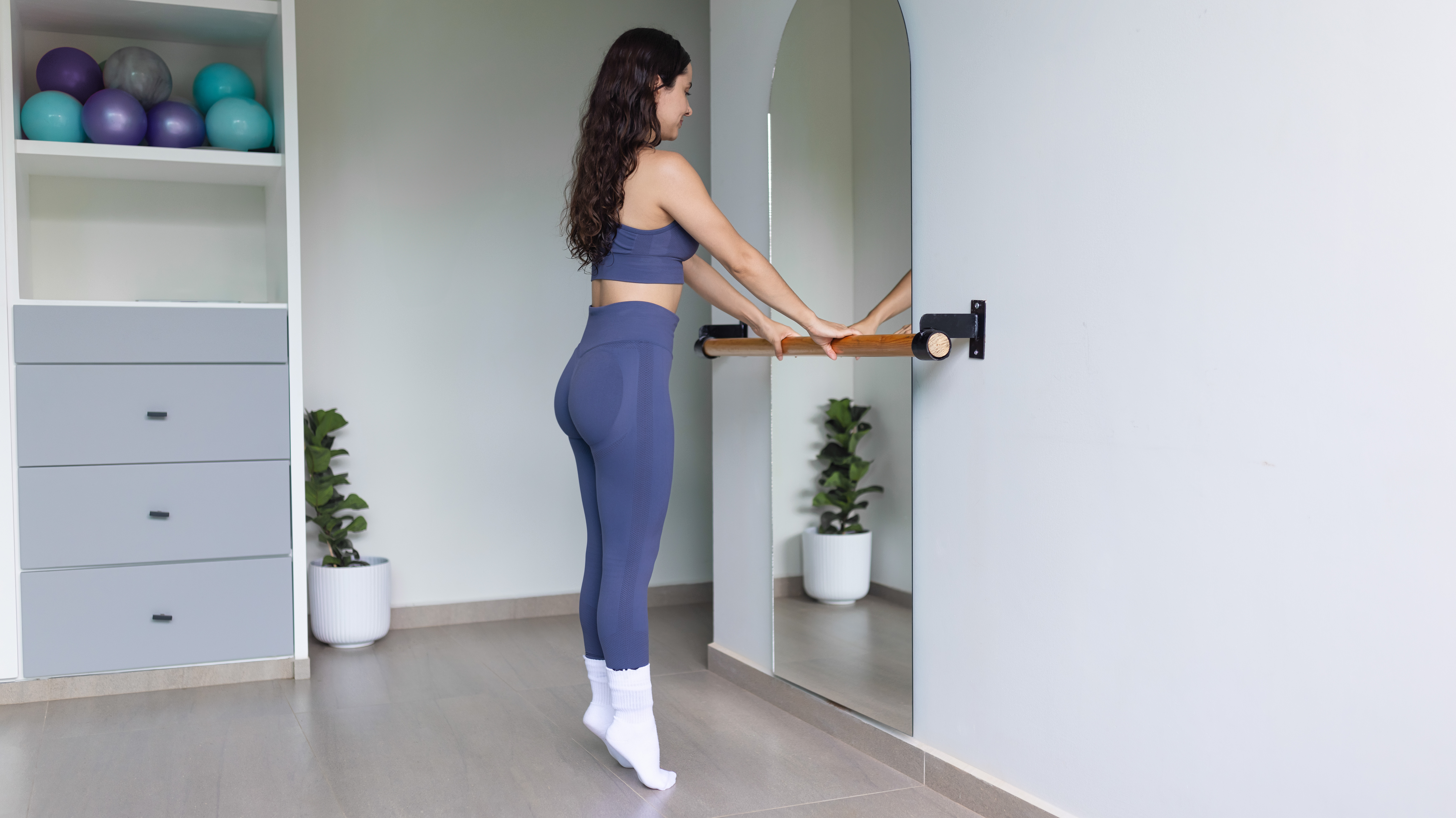 A woman stands at a bar in a studio doing calf raises. She is on her tip toes. In front of her is a mirror, reflecting the green leafy plant behind her. We also see a storage unit, stocked with Pilates balls.