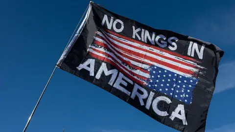 Getty Images A person waves a flag that reads, "NO KINGS IN AMERICA" with the blue sky above it at a Washington DC rally on 17 October - one day before the No Kings protests scheduled in cities across the US. 
