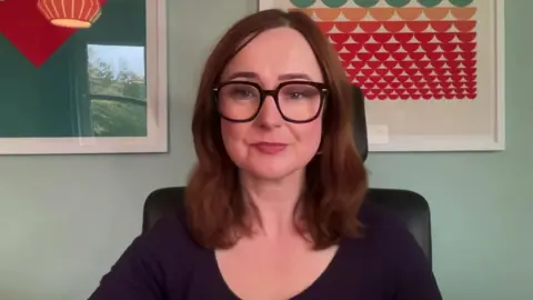 BBC A woman with shoulder length brown hair. She is wearing black framed glasses
She is sat at a desk on a black chair, behind her two colourful prints hang on a pale green painted wall