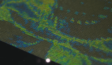 An array of tiny parallel green lines appears over a steel surface. The white dot a laser beam is visible in the lower center of the picture.