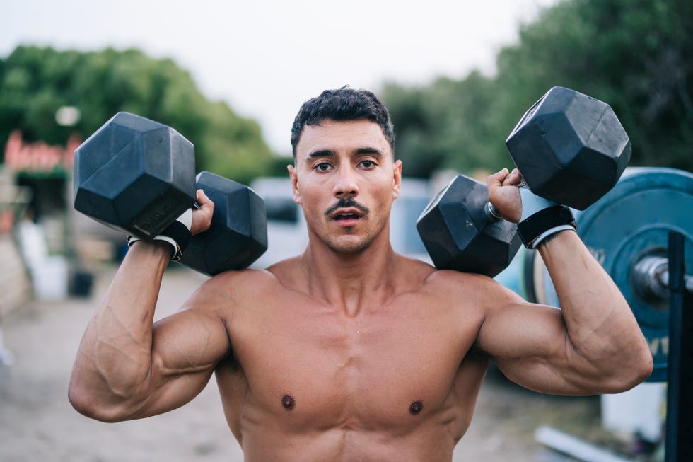 makeshift gym in a courtyard with athletes athletic guy keeping dumbbells on his shoulders looking serious at camera.