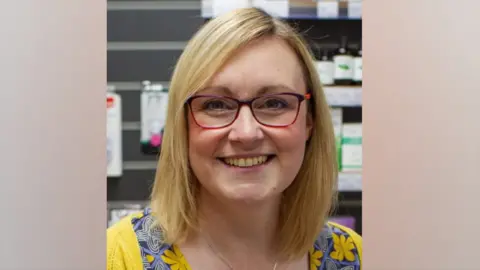 Monkbar Pharmacy A woman with blonde hair and wearing glasses smiles at the camera, in a pharmacy.
