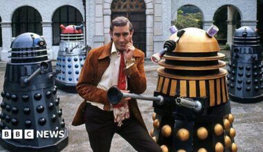 Terry Nation wearing a red tie and tan jacket posing with four multi-coloured Daleks in front of a grand looking building