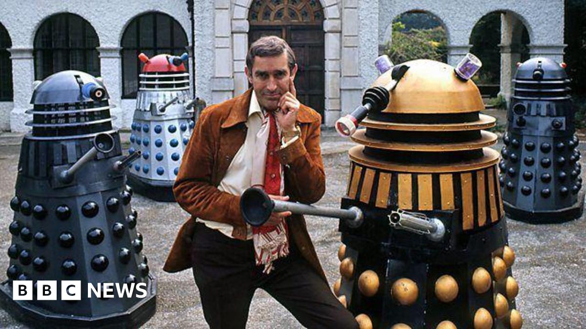 Terry Nation wearing a red tie and tan jacket posing with four multi-coloured Daleks in front of a grand looking building