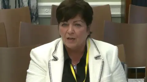 PA Media A head shot of Rita Devlin. She has short brown hair. She is wearing a white blazer with a black shirt. Behind her are rows of brown chairs. She has a yellow lanyard around her neck. 