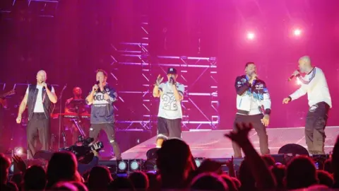 PA Media Five members of the boy band are on a bright pink lighted stage. Each of the members are holding a microphone and appear to be singing. There are people on the bottom of the image watching and a man with a camera filming. 